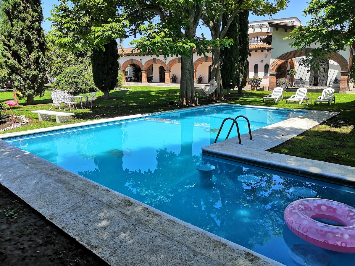 A serene outdoor area features a clear swimming pool bordered by a stone deck. Lush green trees provide shade, and a variety of seating options, including white lounge chairs, are visible. Colorful floaties are resting on the water's surface, while a hammock is positioned nearby.