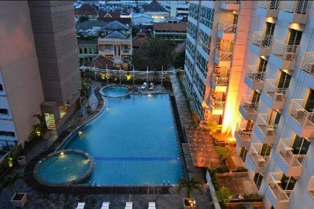 An aerial view captures the large swimming pool, framed by lounge chairs and palm trees. The surrounding buildings are softly illuminated, enhancing the serene atmosphere as evening approaches. The pool area features distinct sections, including a round jacuzzi and a spacious swimming area.