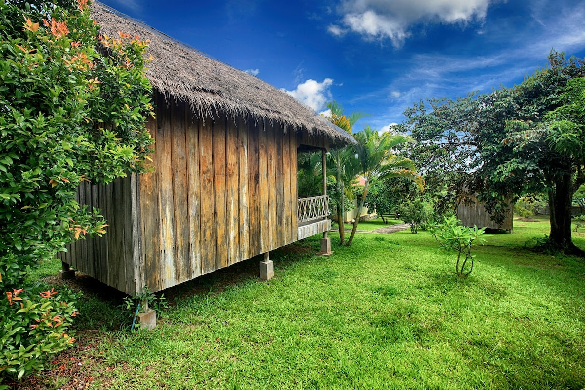 A wooden bungalow features a thatched roof and is elevated on concrete pillars. It is surrounded by lush greenery and colorful shrubs, creating a serene outdoor setting. The sky is partly cloudy, enhancing the natural atmosphere of the area.