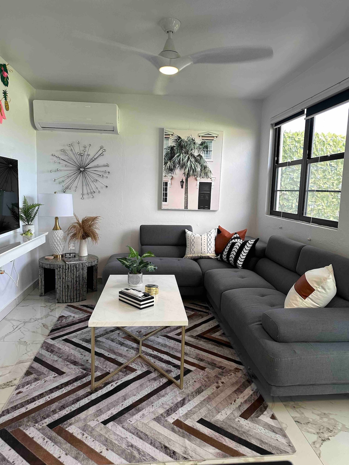 A modern living area features a spacious sectional sofa adorned with various decorative pillows. A stylish coffee table sits atop a geometric patterned rug. Artwork depicting palm trees decorates the wall, while a flat-screen TV is mounted for entertainment. Natural light filters in from the large windows.