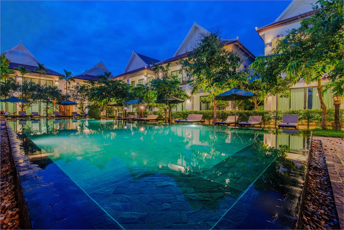 An inviting pool is surrounded by lush greenery and tropical trees, with several sun loungers positioned nearby. The reflections of the buildings are visible on the water's surface, and soft lighting adds a tranquil ambience to the scene.