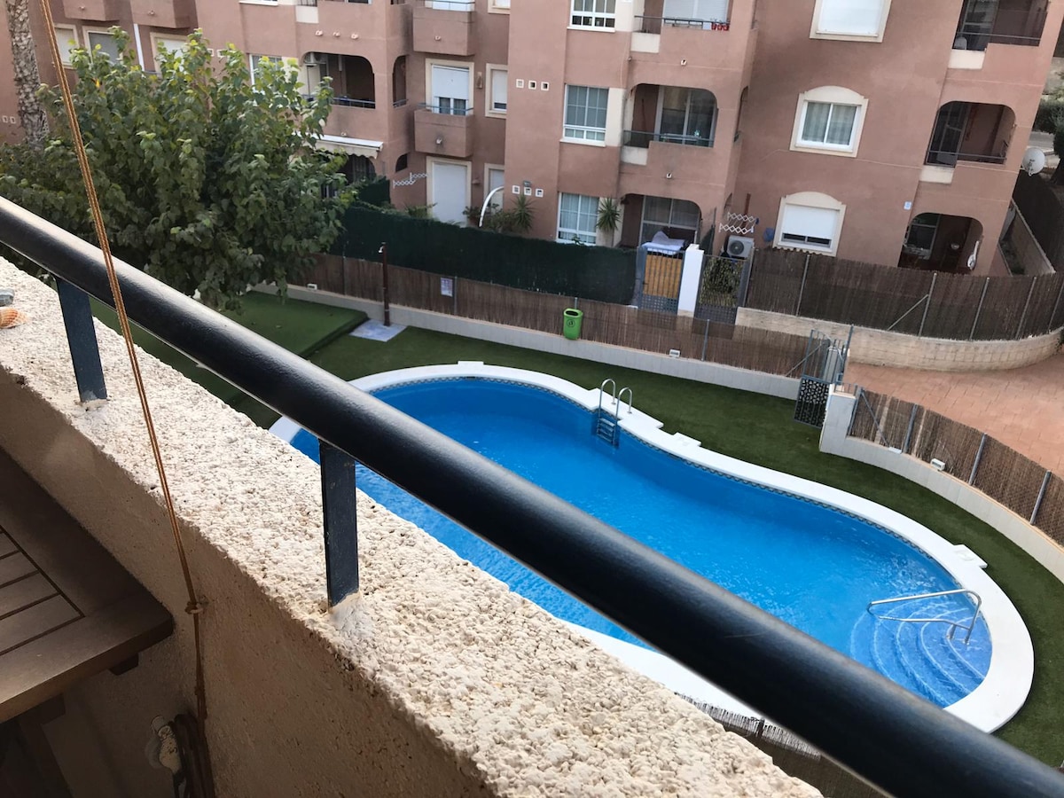 A view from a balcony showcases a curved pool surrounded by well-maintained grass. The pool is framed by low fences, while an apartment complex is visible in the background, providing a tranquil residential setting.