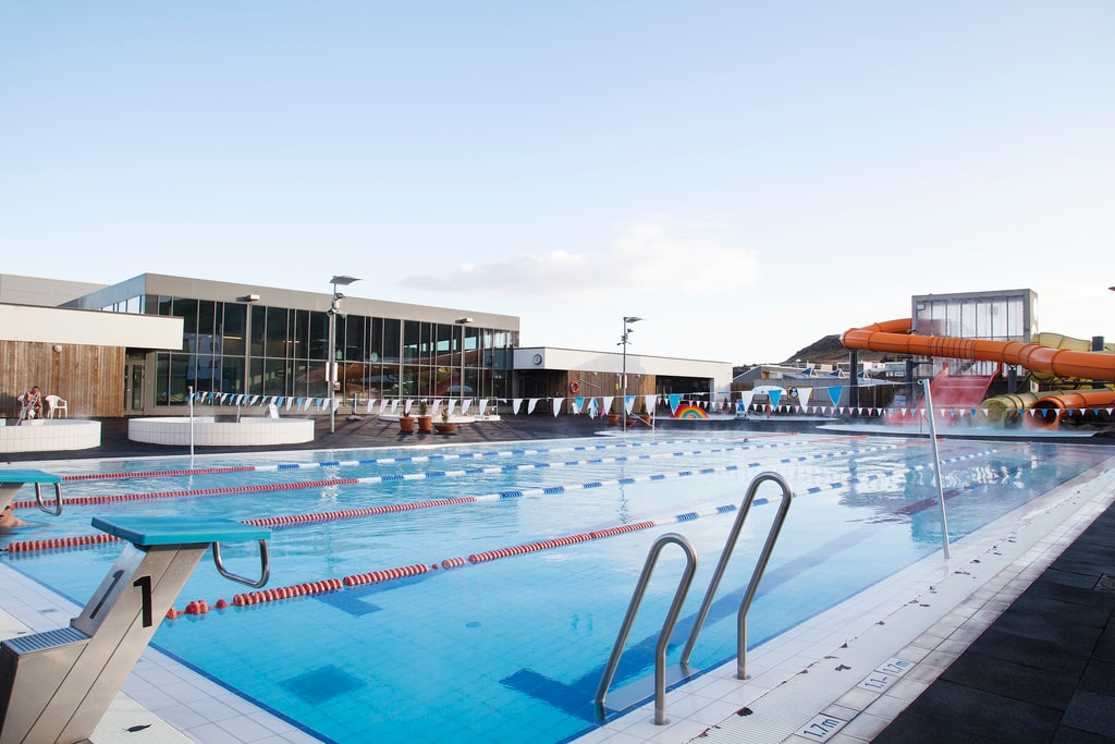 An outdoor swimming pool features multiple lanes and is accompanied by water slides in the background. The area is well-lit and spacious, with sun loungers and diving boards visible near the pool. Clear blue skies are seen above, enhancing the inviting setting.