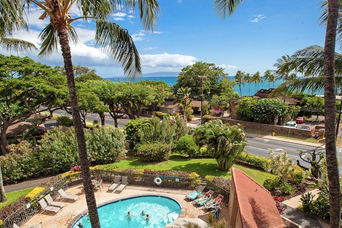 A scenic view features a sparkling pool surrounded by palm trees and lush greenery. In the background, the ocean and blue sky are visible, enhancing the relaxing outdoor atmosphere. Several lounge chairs are placed near the pool, inviting relaxation.