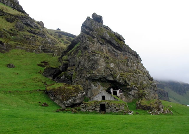 A rustic structure is nestled against a large rocky formation, surrounded by lush green grass. The building features a simple entrance and is complemented by natural stone elements. The landscape showcases a gentle sloping hill, enhancing the serene outdoor environment.