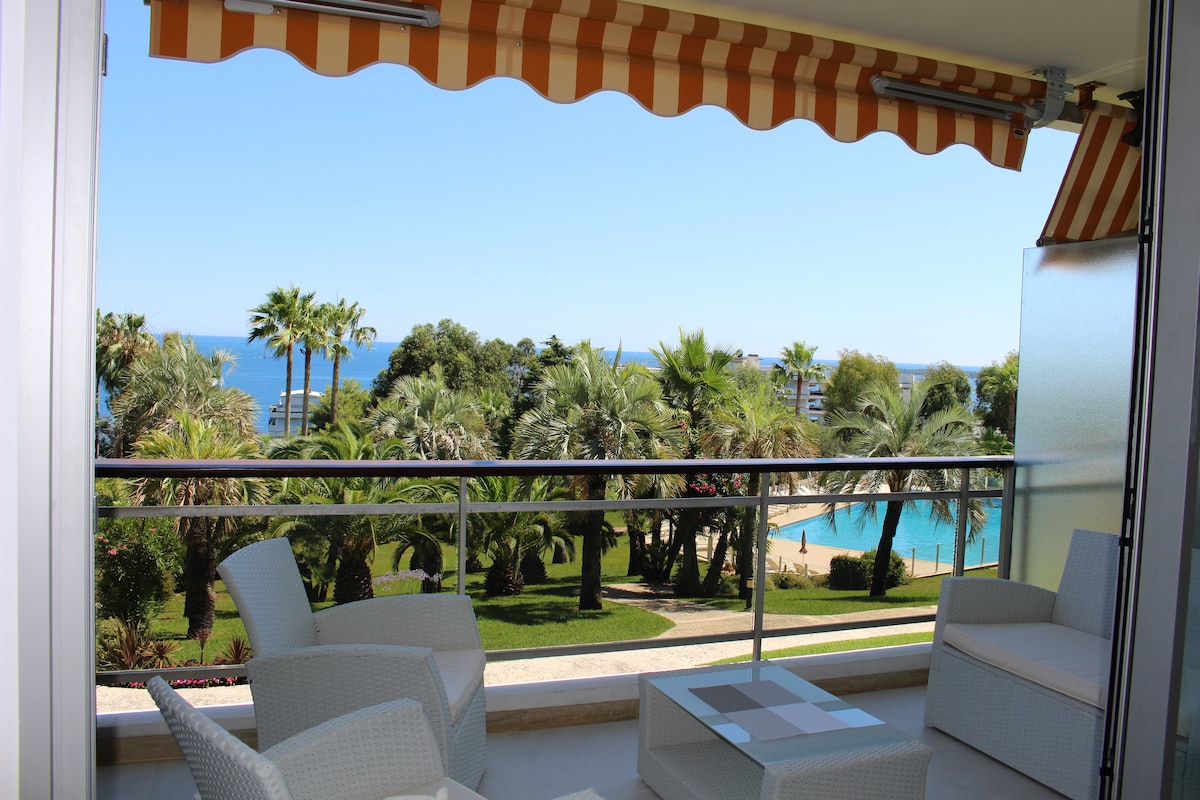 A balcony area is featured, offering a view of the palm trees and the turquoise sea in the distance. Comfortable white chairs surround a small table, with the semi-olympic pool visible below amid lush greenery.