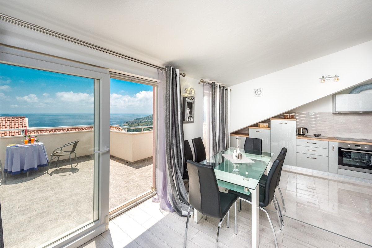 A modern dining area is positioned beside expansive glass doors, providing views of the sea. The space features a sleek glass table surrounded by black chairs. Natural light floods in, and a corner kitchen with contemporary appliances is visible. A shaded outdoor terrace can be seen through the door.