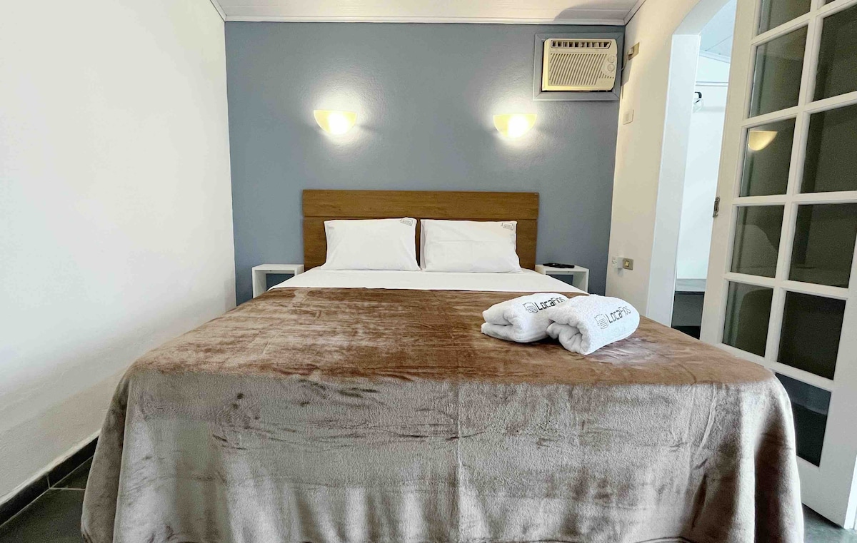 A serene sleeping area is presented with a spacious bed, adorned with a soft brown throw. Two neatly arranged towels rest at the foot of the bed. The room features a light blue accent wall and is equipped with an air conditioning unit on the adjacent wall.