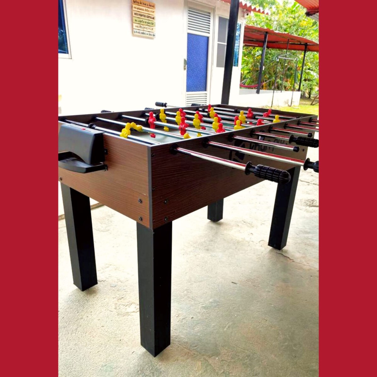 A foosball table features a wooden surface with colorful player figurines in red and yellow. The sturdy black legs provide stability, and the table is positioned on a smooth concrete floor, allowing for easy access and play. Natural light can be seen coming from the background.