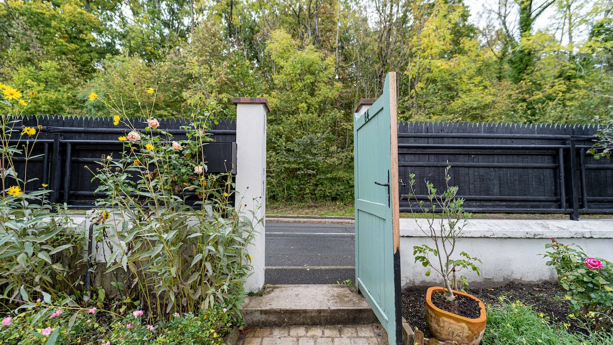 An inviting garden gate opens to reveal a tree-lined pathway bordered by lush greenery. Flowering plants and a decorative pot add a touch of color at the entrance, leading toward a quiet street framed by a wooden fence.