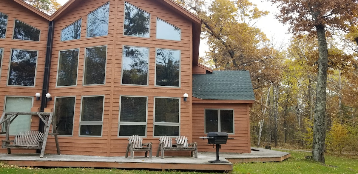 The exterior of the cabin features large windows that fill the space with natural light. A wooden deck houses a gas grill and several seating options, including adirondack chairs. Surrounding trees display autumn foliage, enhancing the cabin's natural setting.