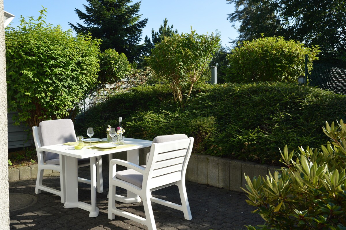 An outdoor seating area is featured, surrounded by greenery. A white table is set with two chairs, suggesting a space for dining or relaxation. Sunlight filters through the foliage, enhancing the natural setting.