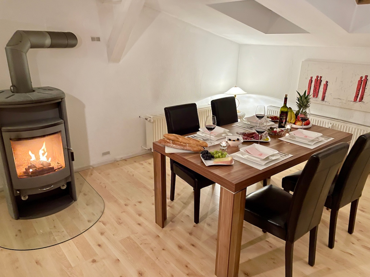 A dining area features a wooden table set for a meal, surrounded by four black chairs. A cozy stove emits a warm glow nearby. The table is arranged with plates, glasses, and a variety of dishes, complemented by a subtle light from a nearby lamp.