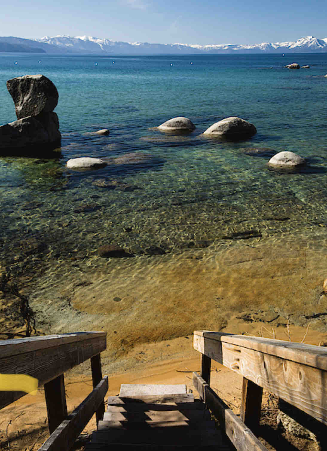 A wooden staircase descends towards a clear lake, surrounded by smooth boulders and small pebbles. The tranquil water reflects the sky and distant mountains, creating a serene natural landscape. Sunlight enhances the golden sandy beach visible at the base of the stairs.