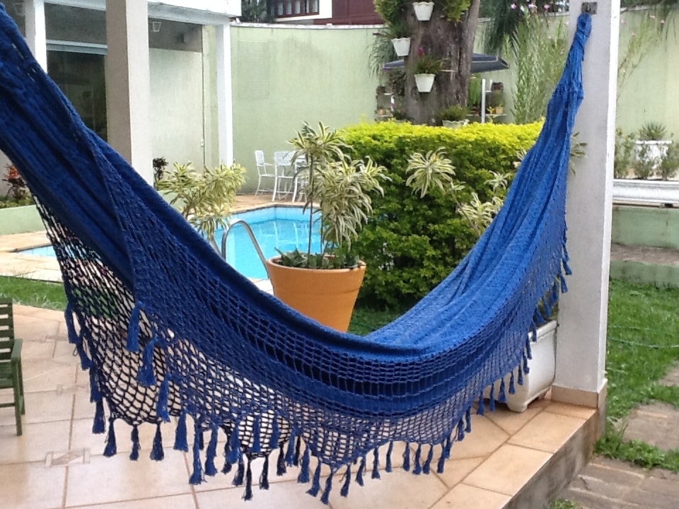A blue hammock is strung between two posts, providing a relaxing spot near a swimming pool. Surrounding greenery and potted plants create a lush, calming environment. The tiled floor offers a spacious area for outdoor enjoyment.