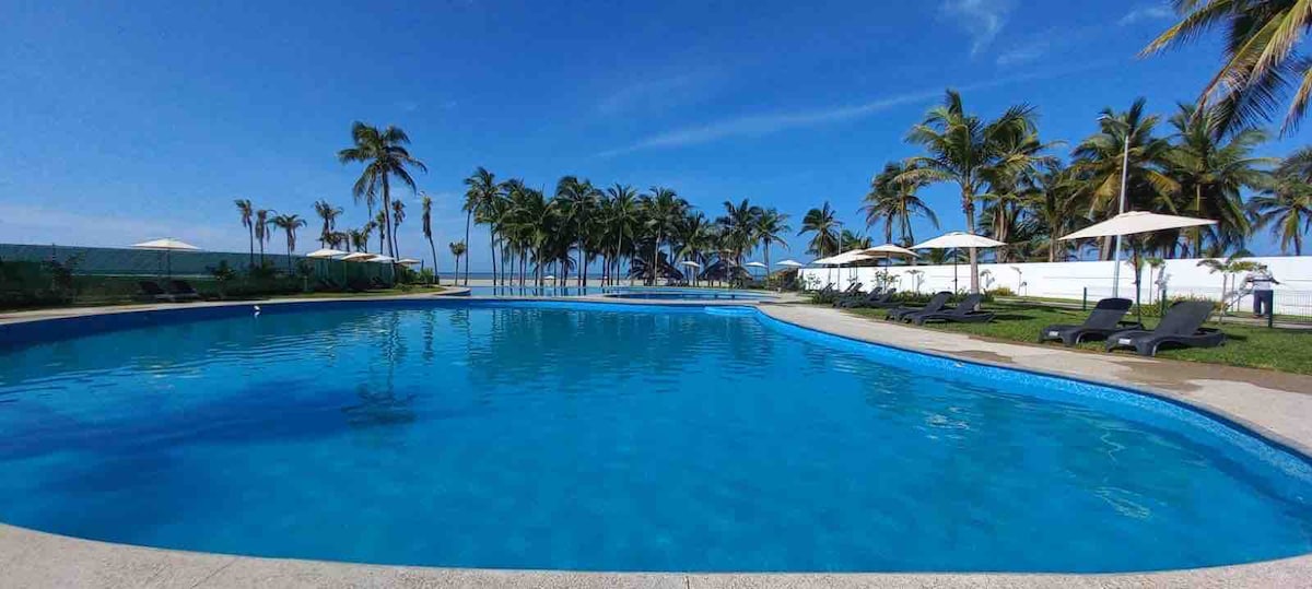 A large swimming pool reflects the bright blue sky, surrounded by well-maintained green lawns and palm trees. Comfortable lounge chairs are strategically placed along the pool's edge, providing a relaxing spot for guests to enjoy the tropical setting.
