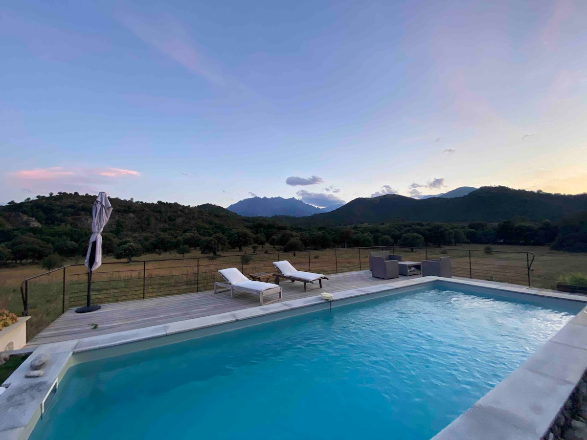 A private heated pool is set against a backdrop of rolling hills and distant mountains. Two sun loungers are positioned on the wooden deck, inviting relaxation. The tranquil scene is enhanced by a soft sunset glow, reflecting off the clear water of the pool.