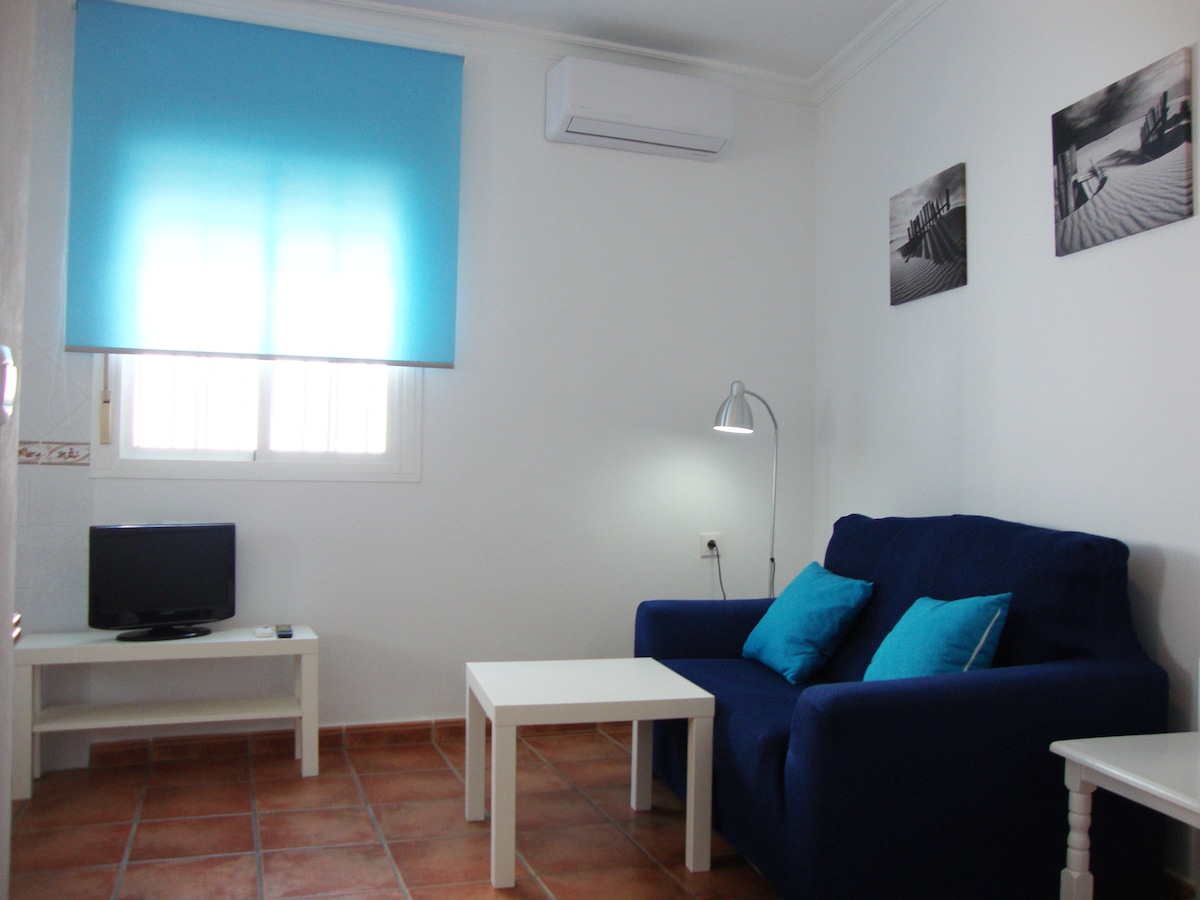 A cozy living area features a blue sofa with decorative cushions, positioned near a small white coffee table. A wall-mounted television is present, along with a floor lamp providing additional light. A large window is covered by a blue shade, allowing natural light to enter.