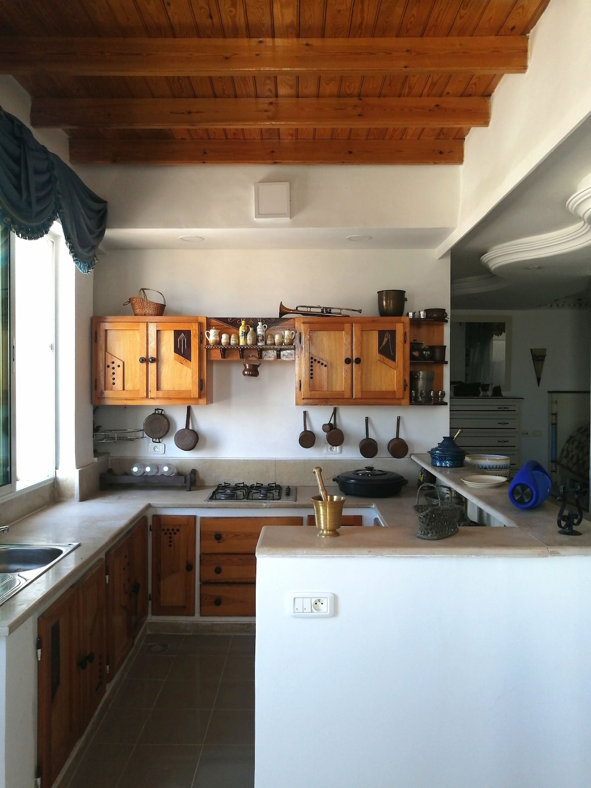 A compact kitchen area features wooden cabinetry with decorative accents and an array of cooking utensils displayed on the wall. A gas stove and sink are integrated into the countertop, while natural light enters through a window draped with navy blue fabric.