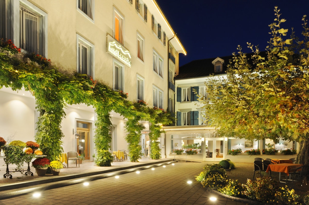 The entrance of the hotel is adorned with greenery and seasonal flowers, providing a lively contrast to the soft lighting. Tables and chairs are arranged along the outdoor area, welcoming guests to relax in a tranquil environment.