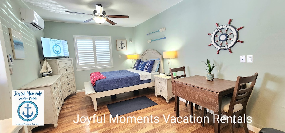 A cozy studio room features a king bed with light-colored bedding, complemented by wood flooring. A small dining table sits near a window, enhancing the natural light. Decorative nautical elements, including a ship's wheel and an anchor, adorn the walls, creating a relaxed atmosphere.