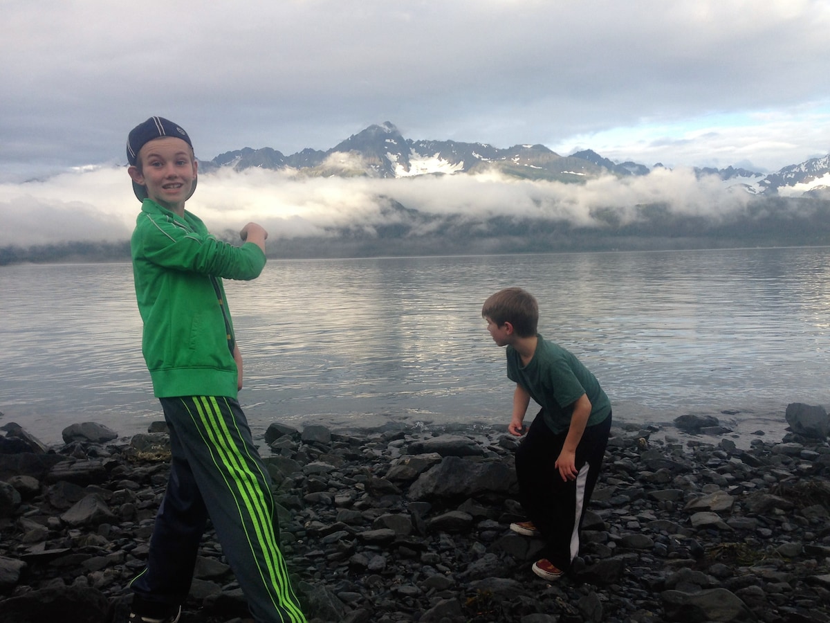 Two children are seen playing by a rocky shoreline, with calm waters reflecting the surrounding mountains and cloudy sky. The landscape features lush greenery, and the serenity of the scene is enhanced by the natural beauty of the Alaskan environment.