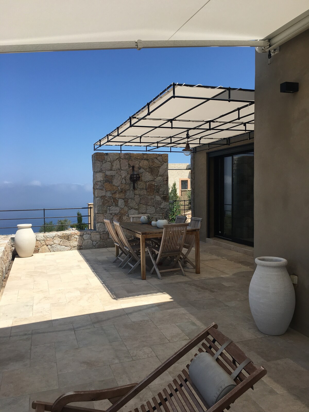 An outdoor dining area is showcased, featuring a sturdy wooden table surrounded by sleek chairs. Large planters and stone walls enhance the space. A retractable awning provides shade, while expansive views of the horizon are visible in the background.