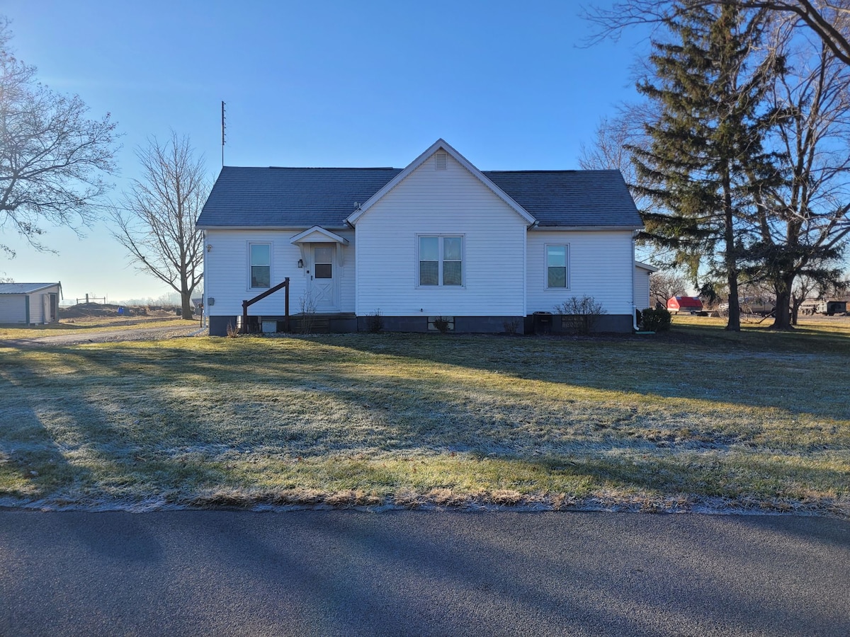 A simple, one-story house features a light-colored facade with a pitched roof and front steps. Flanked by two small windows, the front door is centrally located. A large grassy area surrounds the home, with trees and open space visible in the background under a clear blue sky.