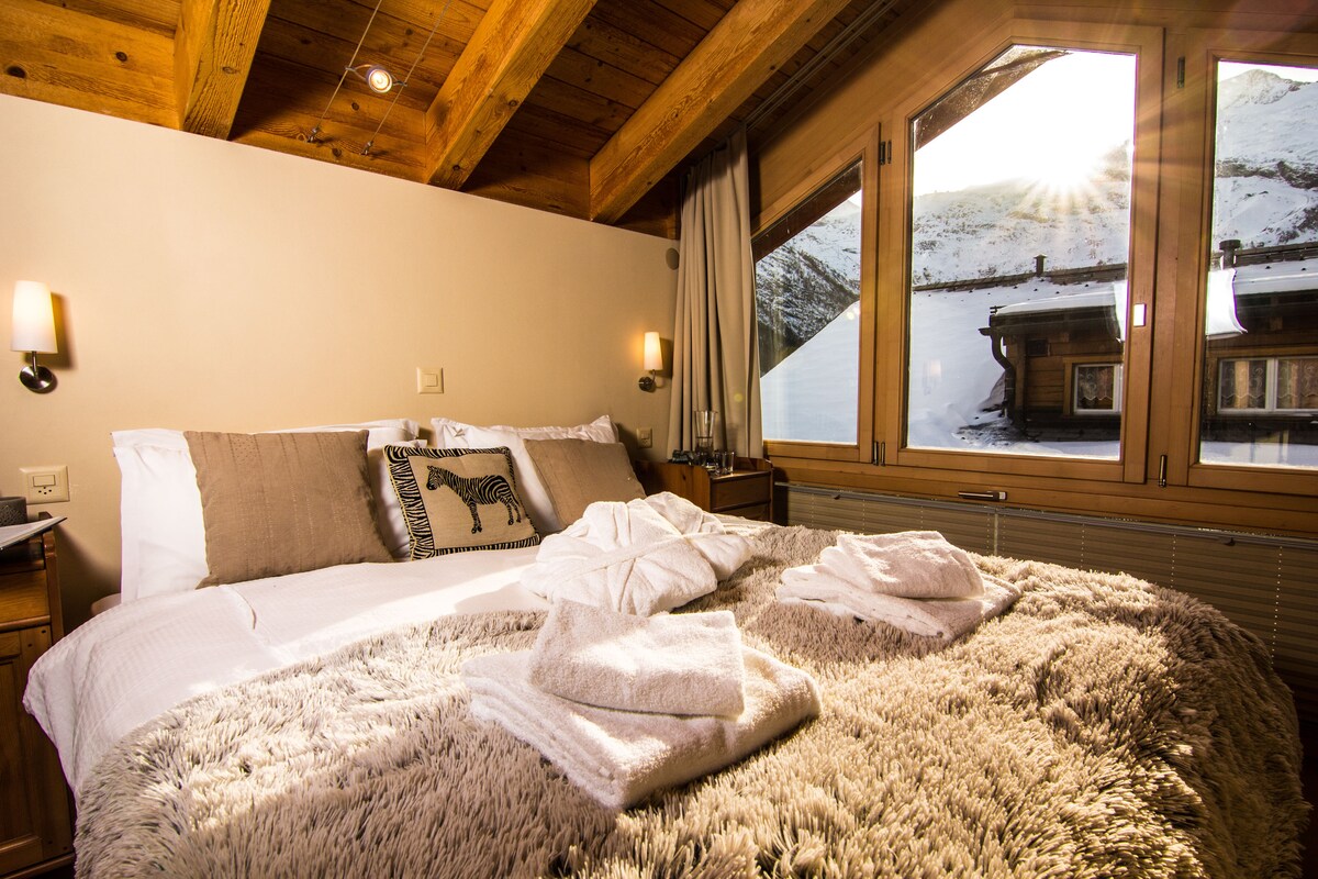 A spacious bedroom features a large bed dressed in soft linens and accompanied by plush, folded towels. Natural light streams in through large windows, highlighting wooden beams in the ceiling and offering views of the snowy exterior.