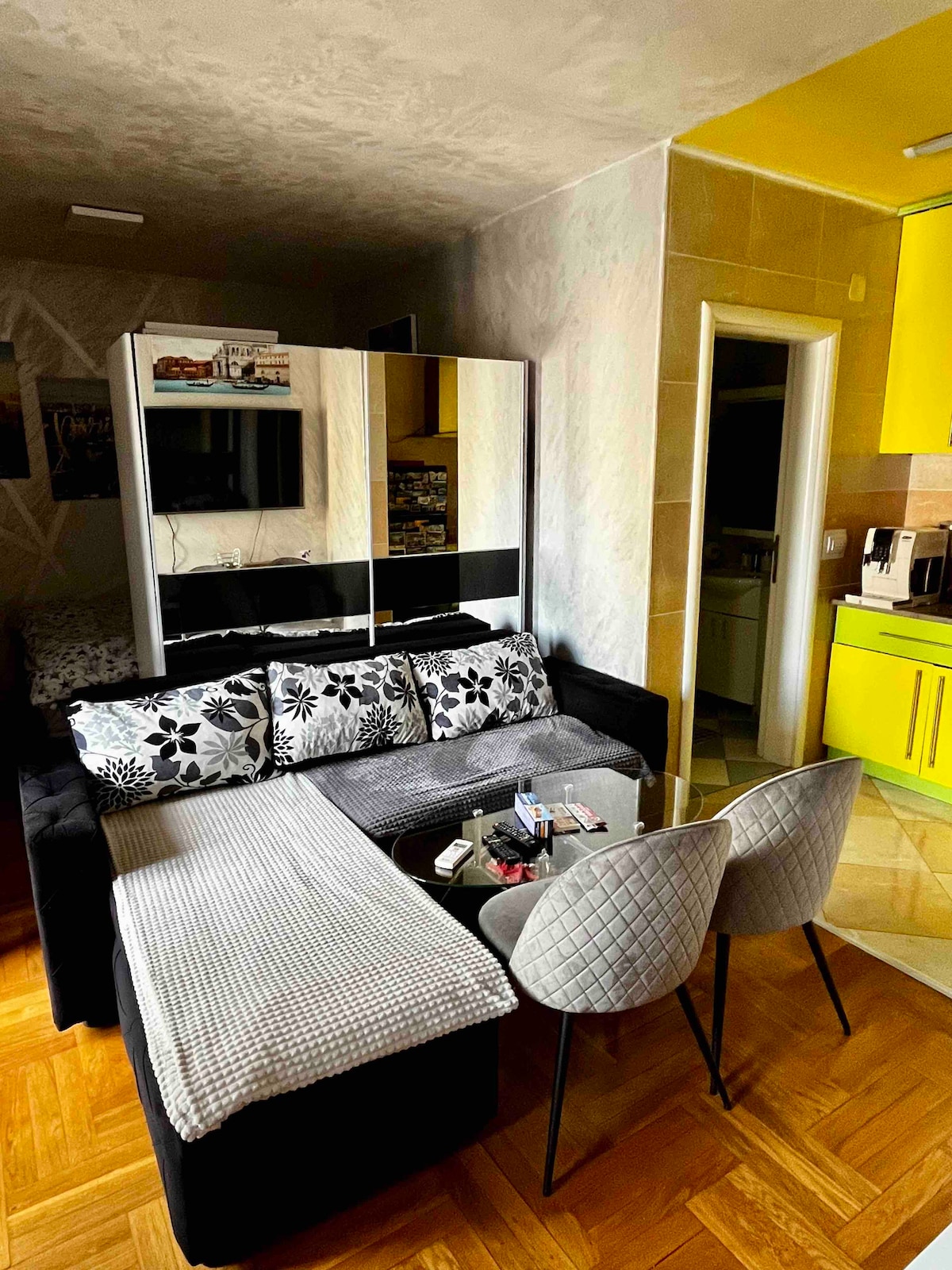 A cozy living area is presented with a dark sofa adorned with floral cushions. A glass coffee table is positioned in front of the sofa, while two light gray chairs provide seating. Bright yellow cabinetry can be seen in the background, complementing the overall warm ambiance.