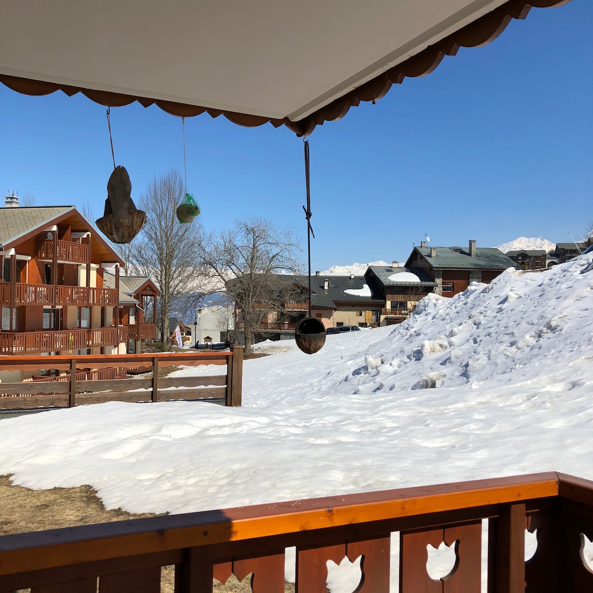 A view from the balcony showcases snow-covered ground and distant mountains under a clear blue sky. Wooden structures with balconies are visible in the foreground, creating a sense of the charming landscape surrounding the apartment.