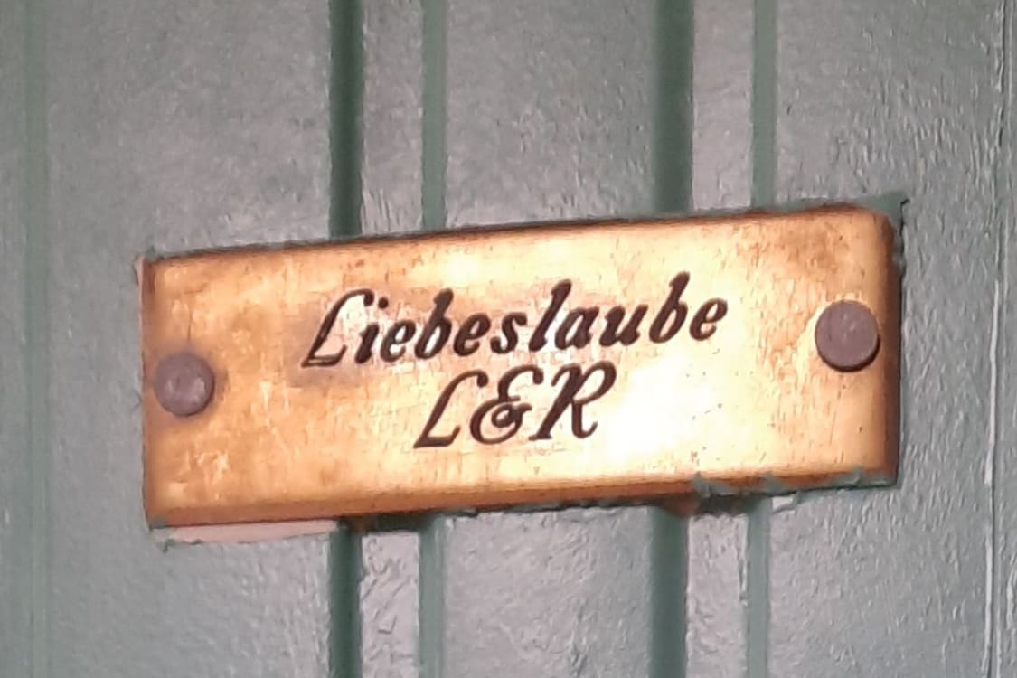 A brass sign with the inscription 'Liebeslaube L&R' is mounted on a wooden panel. The sign’s warm tone contrasts with the vertical green stripes of the wooden surface behind it.
