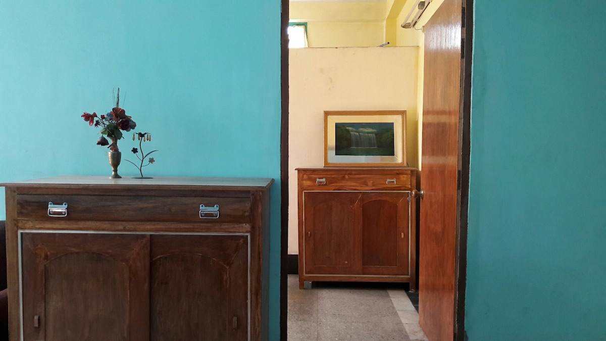 An entrance area features two wooden cabinets against a turquoise wall. A small vase with dried flowers is displayed on the first cabinet. A doorway leads to a light-colored room, where a framed landscape artwork is visible on a side cabinet.
