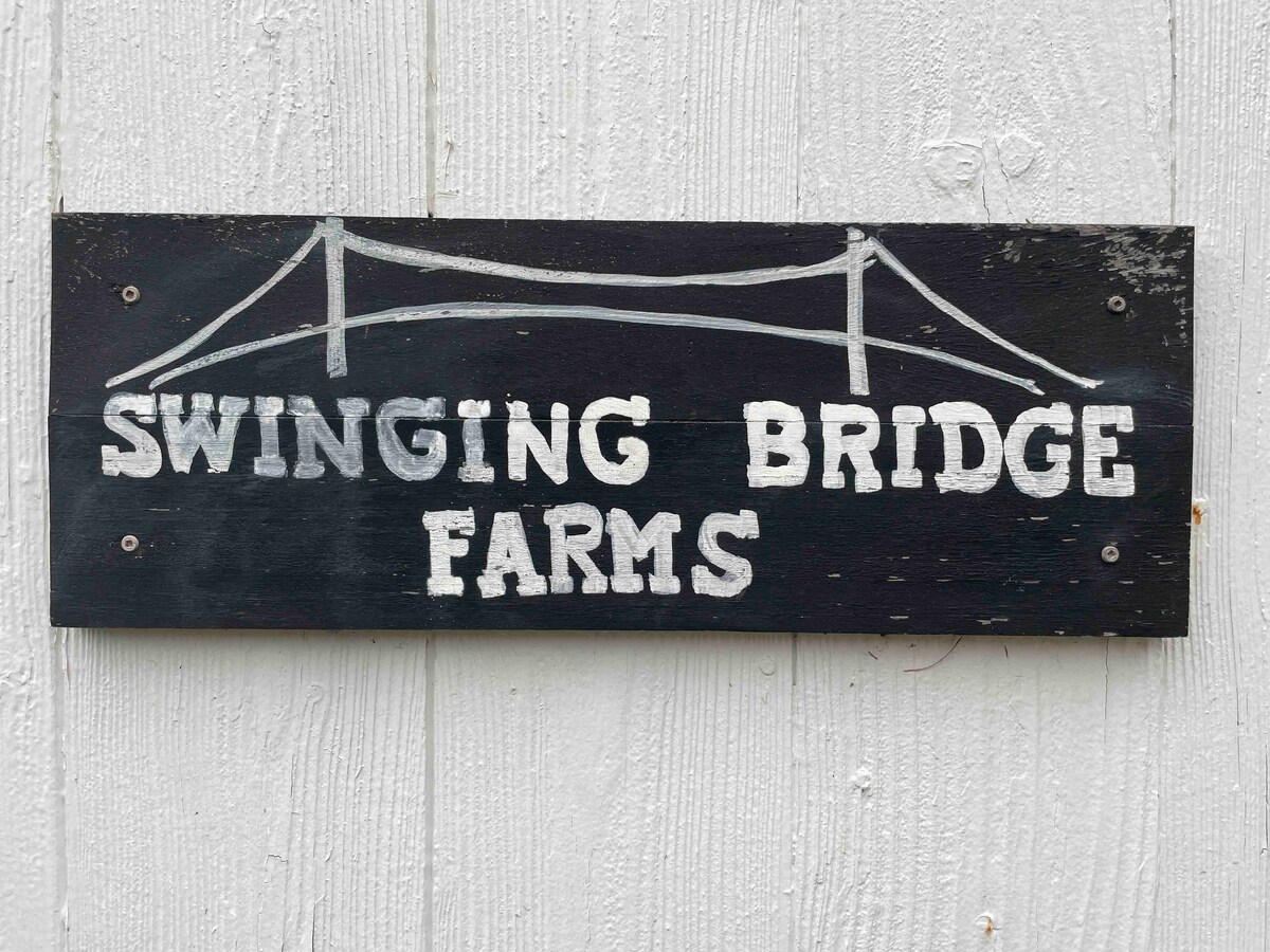 A wooden sign is displayed on a white wall, indicating 'SWINGING BRIDGE FARMS' in bold, white lettering. The sign features a simple illustration of a swinging bridge above the text, enhancing the rustic charm of the farm setting.