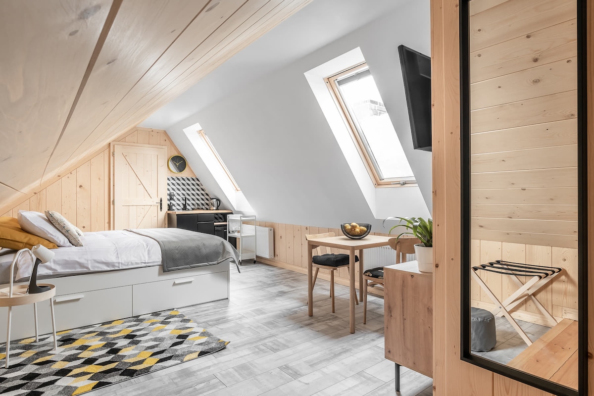 A bright loft space features angled ceilings and large skylight windows, allowing ample natural light. A cozy bed with neutral bedding is positioned beside a small dining area, which includes a table and chairs. Wooden accents create a warm, inviting environment.