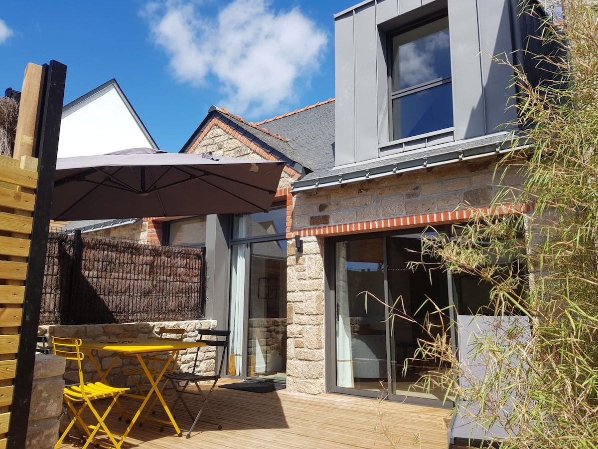 An outdoor terrace features a small yellow table and chairs, shaded by a large umbrella. The stone and modern materials of the villa's exterior create a balanced look, with large glass doors providing access to the interior, framed by a backdrop of greenery.