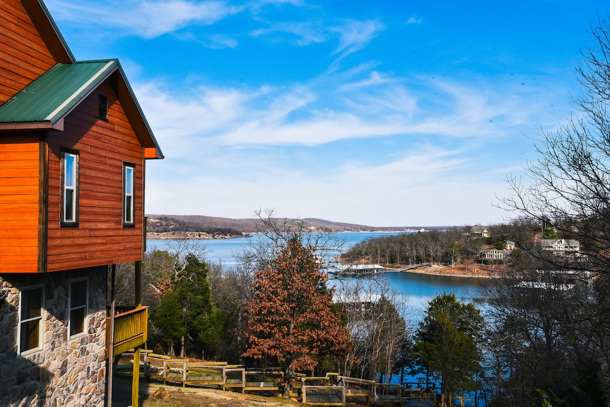 The lodge exterior is presented against a clear blue sky, showcasing a blend of stone and wood siding. The scenic view highlights a serene lake and wooded surroundings, complemented by a gentle slope leading down to the water.