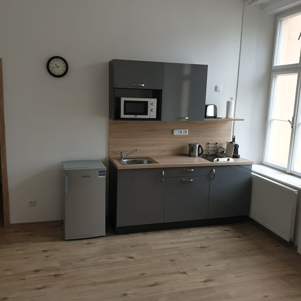 A modern kitchen area is presented with sleek gray cabinetry and wooden countertops. A microwave and kettle are positioned on the counter, alongside a coffee maker. A compact refrigerator stands nearby, with natural light streaming in from the window, highlighting the clean and functional design.