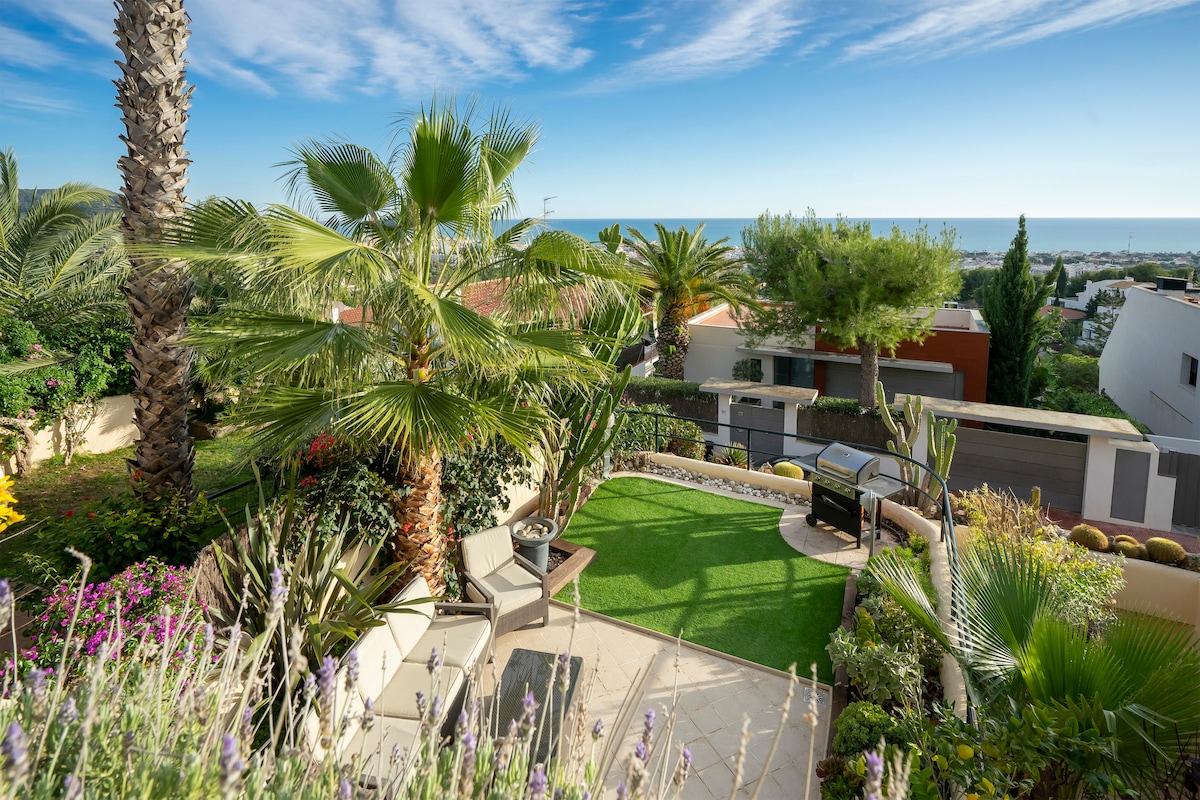 A garden area is surrounded by lush palm trees and colorful flowers, featuring a grassy space with comfortable seating and a barbecue. The backdrop showcases a serene view of the sea and neighboring homes under a clear blue sky.