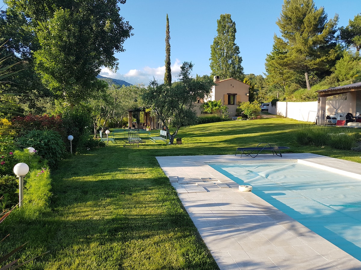 A spacious garden features a well-maintained lawn and a swimming pool surrounded by lounge chairs. Mature trees provide shade, while a house is positioned in the background. Outdoor seating can be seen under a covered terrace, creating a tranquil outdoor environment.