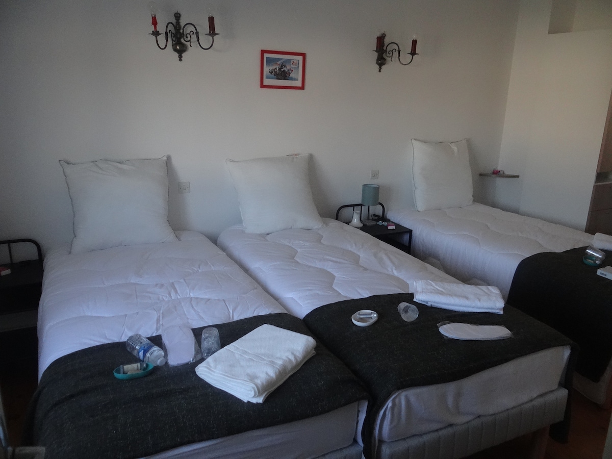 A sleeping area is arranged with three individual beds, each made with white linens and oversized headboards. Dark bedding contrasts with the light walls, while bedside tables hold small lamps. Wall sconces provide additional lighting, and framed artwork adds a touch of decor.