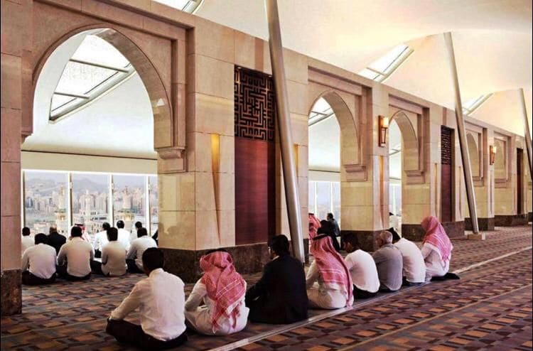 The image shows a spacious prayer room featuring large windows that offer a view of Makkah city. A group of individuals is seated on the floor, adorned in traditional attire, while architectural arches and modern pillars enhance the serene atmosphere. Natural light fills the area.