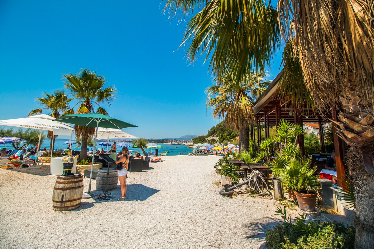 A sandy beach area is framed by palm trees and umbrellas, providing shaded spots. In the foreground, a person stands near a barbecue setup. Beach loungers and umbrellas dot the sandy landscape, with a view of the clear blue water in the background.