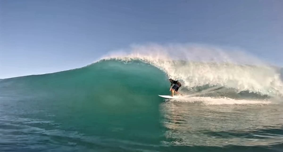 A surfer rides a large, curling wave, showcasing the fluid motion of water. Sunlight reflects off the wave's surface, creating a vibrant turquoise hue. The ocean's texture appears smooth, capturing the dynamic energy of the surf.