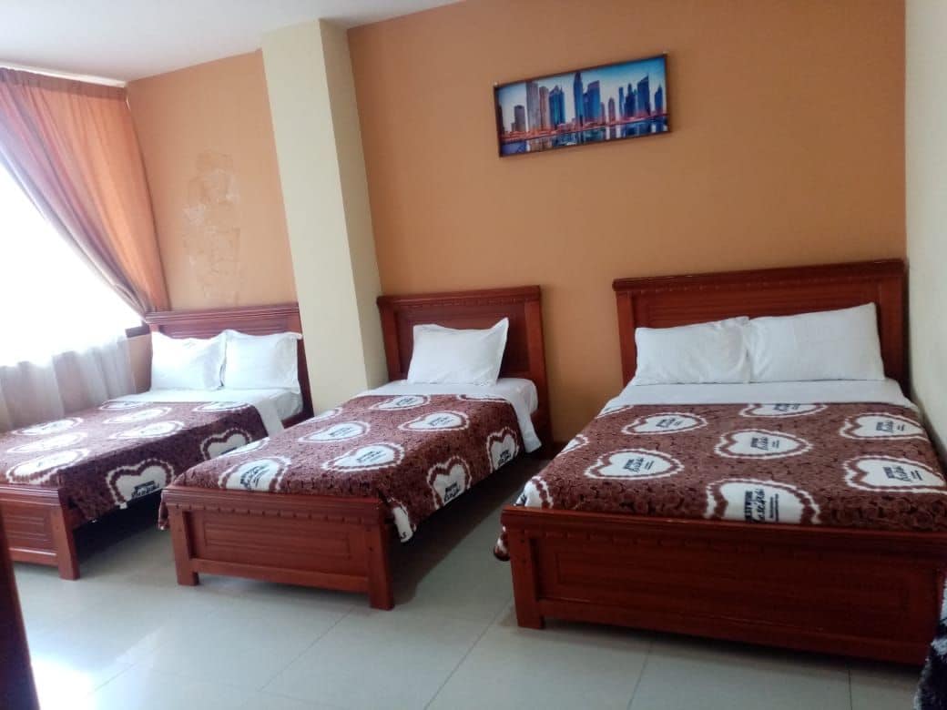 A bright guest room is furnished with three spacious beds, each topped with patterned brown blankets. Soft light enters through a large window, complemented by sheer curtains. A framed cityscape artwork is displayed on the wall, adding a touch of urban charm.