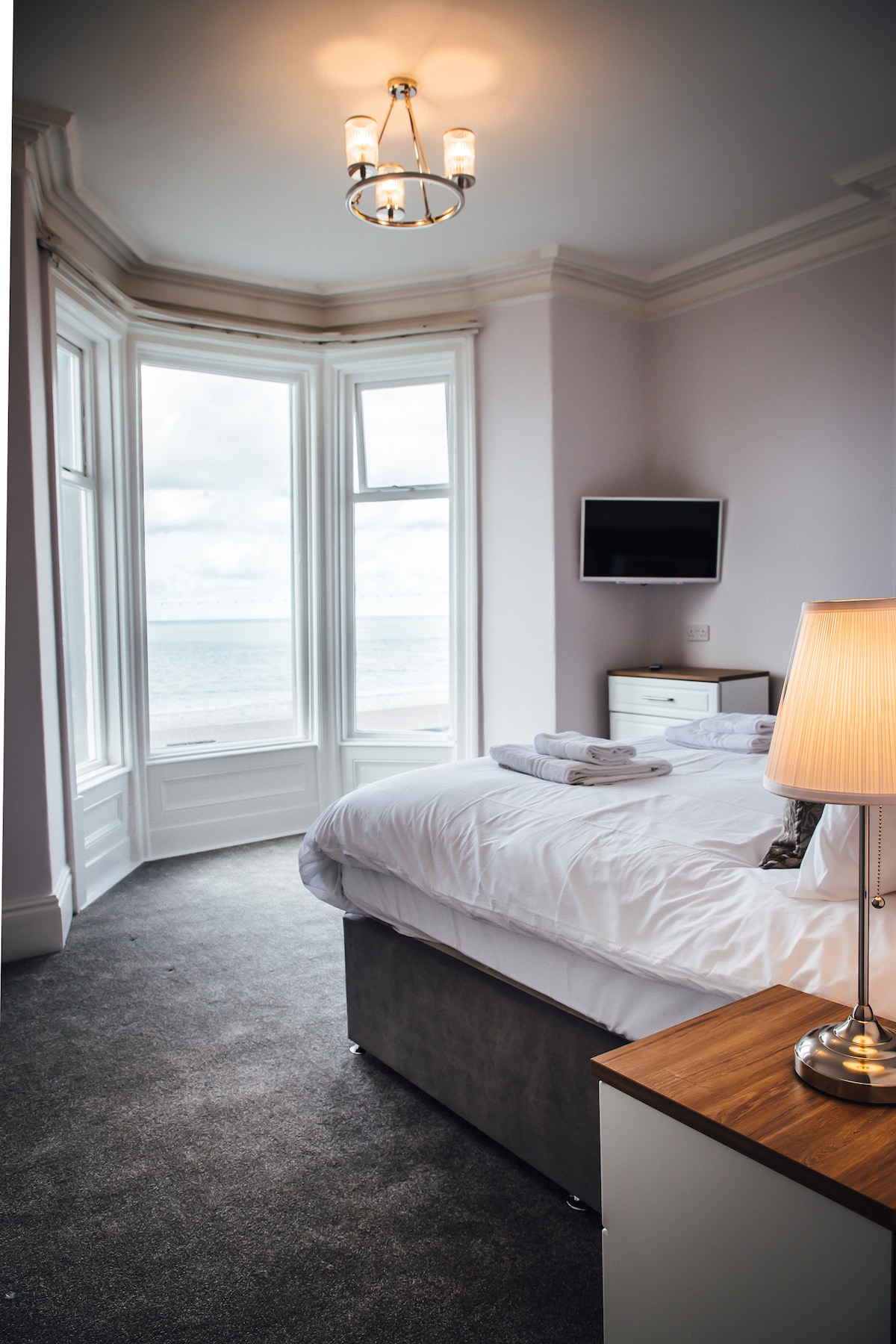 A light-filled bedroom features large bay windows offering views of the sea. A spacious bed is neatly made with fresh linens, and towels are arranged on top. A bedside lamp and a small television are also visible, complementing the modern décor.