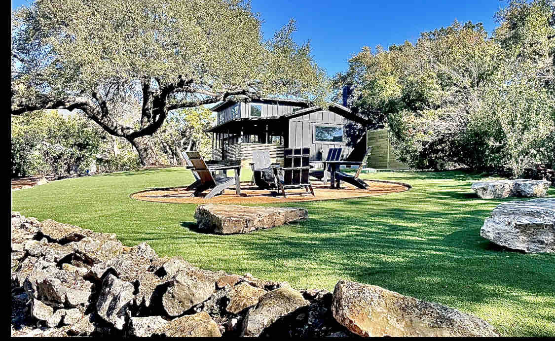 A spacious outdoor area features lush green grass, surrounded by natural stone borders and large rocks. Comfortable seating arrangements are positioned around a central fire pit, with a quaint black house visible in the background against a backdrop of trees.