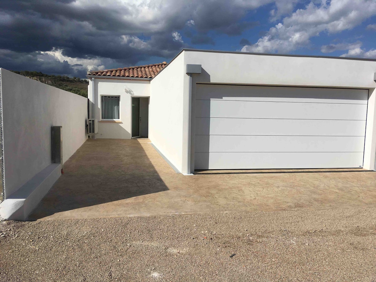 A modern, single-story home is depicted, featuring a sleek garage door and a minimalistic exterior. The entrance is accessed via a paved pathway, while a window and an air conditioning unit are visible on the side of the house, surrounded by a gravel driveway.