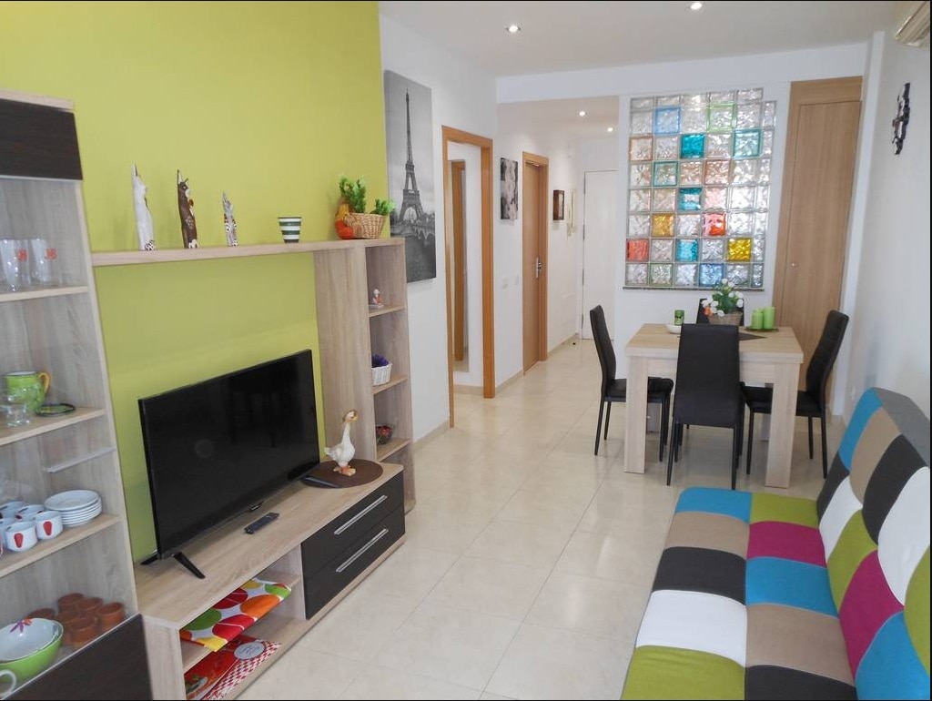 A living area is showcased with a colorful sofa, complemented by a wooden shelf containing decorative items and a television. A dining table with chairs is visible in the background, alongside a wall adorned with a colorful glass block window, enhancing the room's design.