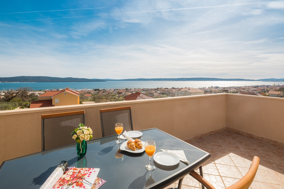 A balcony table is set for breakfast, featuring orange juice and croissants. A vase of flowers adds a touch of elegance. The expansive view showcases the Adriatic Sea and coastal landscape under clear blue skies.
